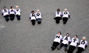 Women lie on the ground wearing white t-shirts with a letter on the front. Lying together they spell out "SAY NO TO NATO"