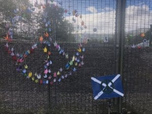 Peace cranes arranged on a fence to form the image of the CND symbol. A home made placard of a Scottish flag with CND symbol in the middle stands beside it.
