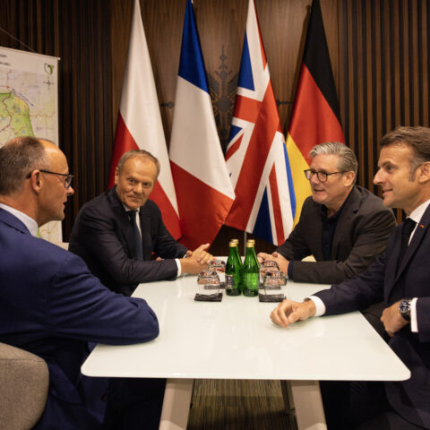 Prime Minister Keir Starmer, Emmanuel Macron, French President, and Friedrich Merz, German Chancellor and Poland Prime Minister Donald Tusk sit ay a table with flags of their respective countries in the background.
